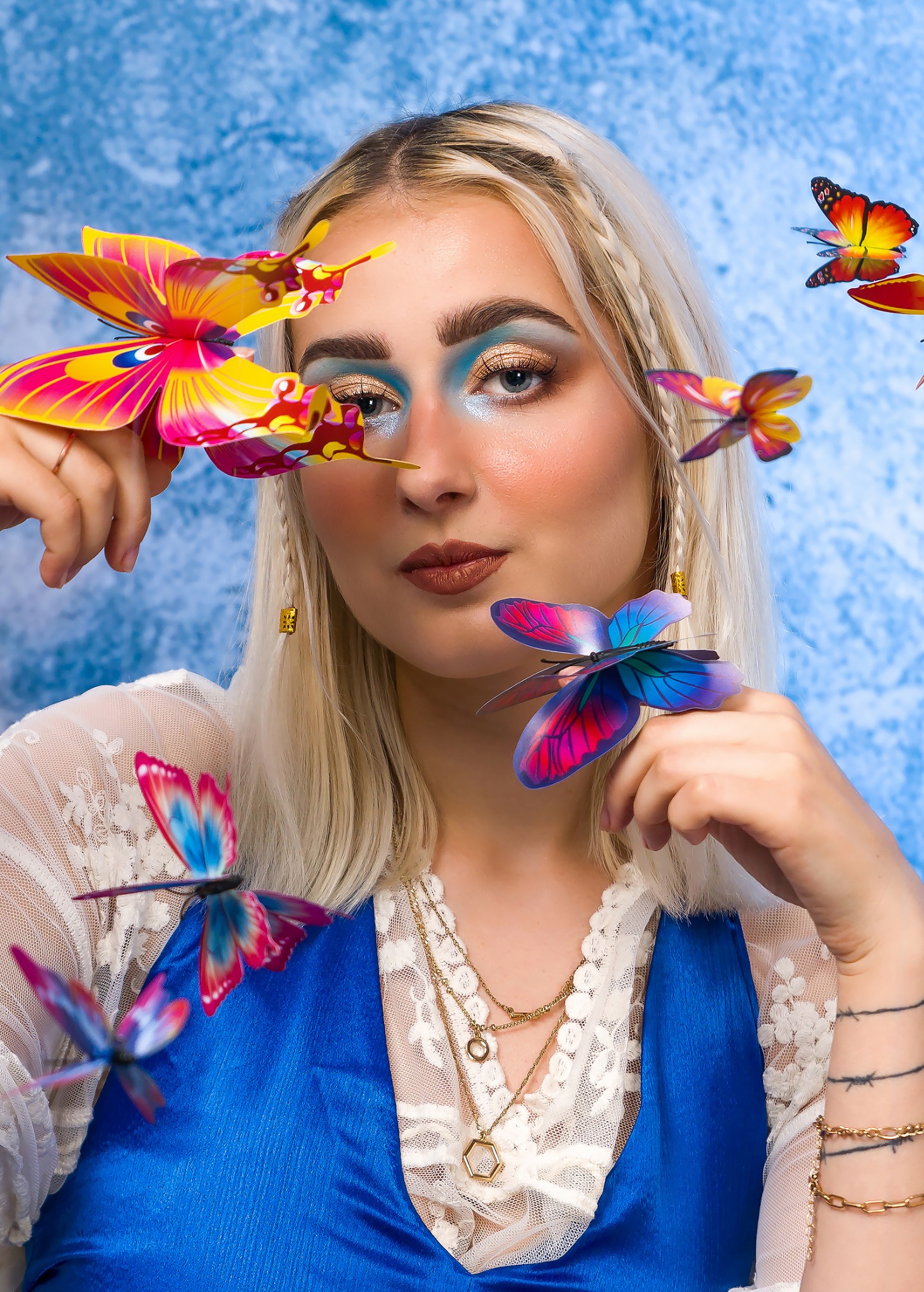 Portret kobiety z blond warkoczami, w niebieskim gorsecie i koronkowej bluzce, otoczonej kolorowymi papierowymi motylami, z intensywnym, błyszczącym makijażem oczu.