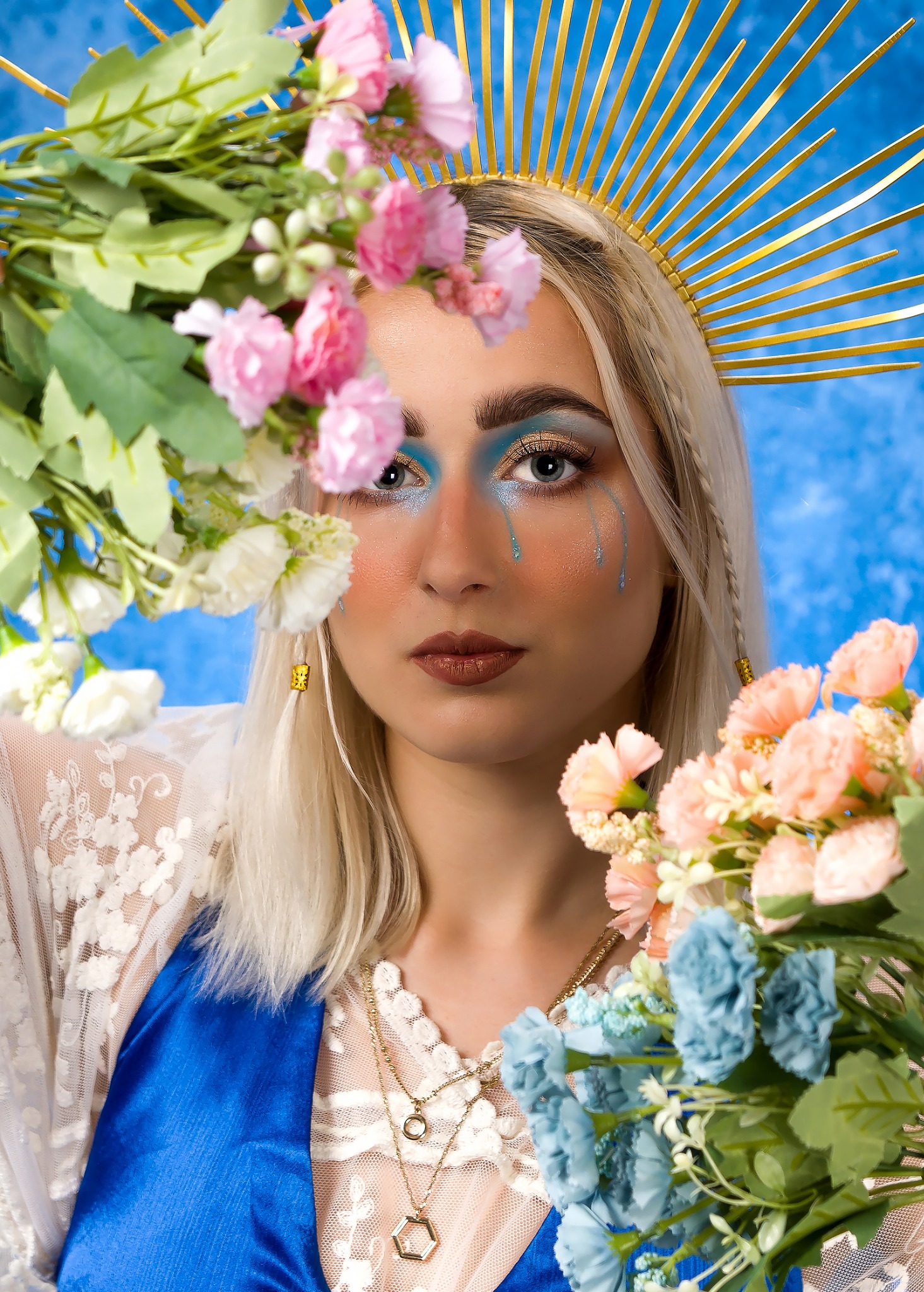 Portret kobiety z blond warkoczami, w koronie ze złotych promieni, z niebieskim makijażem i ozdobnymi łzami, otoczonej bukietami różowych i niebieskich kwiatów na tle błękitnego nieba.