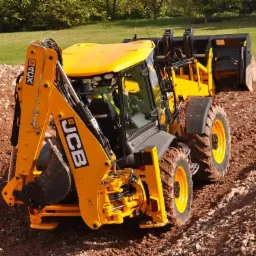 Żółta koparko-ładowarka JCB na nierównym terenie z pryzmą ziemi w tle, widok z tyłu.