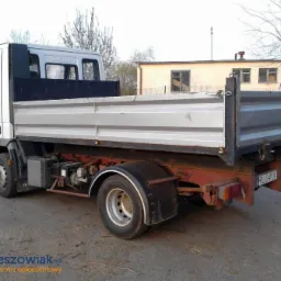 Biała wywrotka Iveco z aluminiową skrzynią ładunkową, widoczna z boku na asfaltowym placu, z budynkiem w tle.