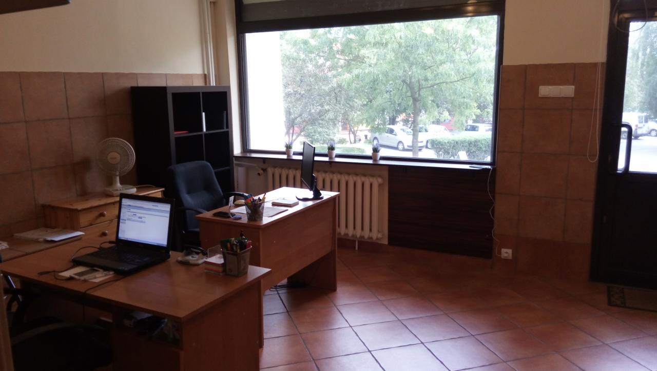 Biuro z dwoma biurkami, komputerem, monitorem, wentylatorem, regałem i oknem z widokiem na zieleń.