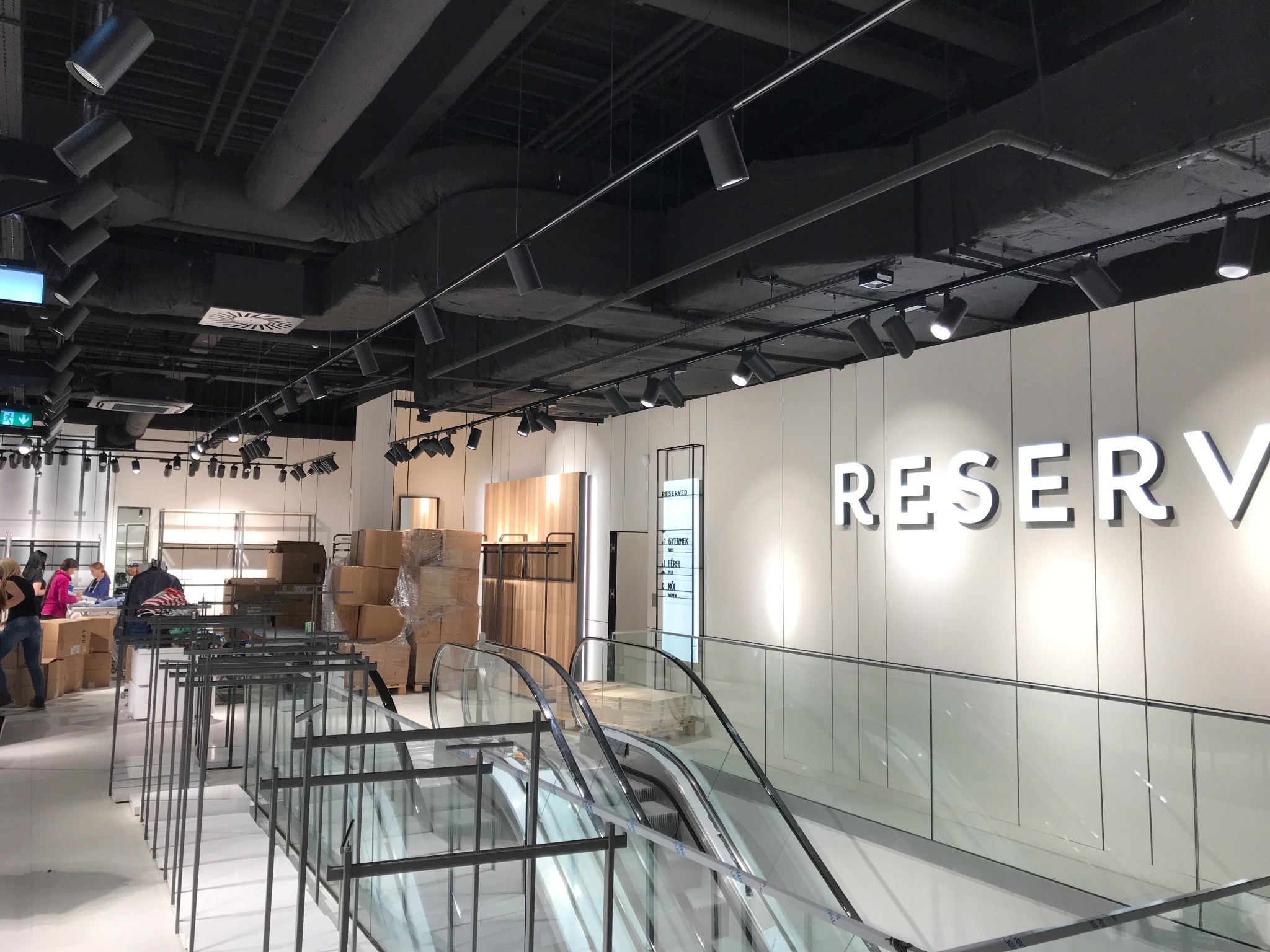 Wnętrze sklepu odzieżowego w trakcie aranżacji: widoczne kartony z towarem, metalowe ramy ekspozycyjne, schody ruchome i oświetlenie punktowe na czarnym suficie.