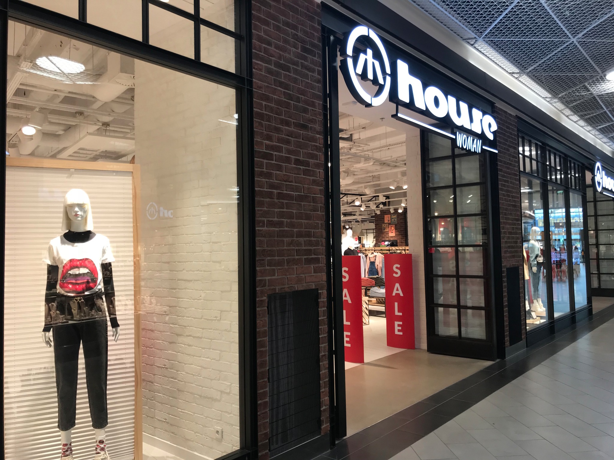 Elewacja sklepu odzieżowego z cegły klinkierowej, czarne ramy okienne, witryna z manekinem prezentującym ubrania z nadrukiem ust, czerwone szyldy z napisem SALE przy wejściu.