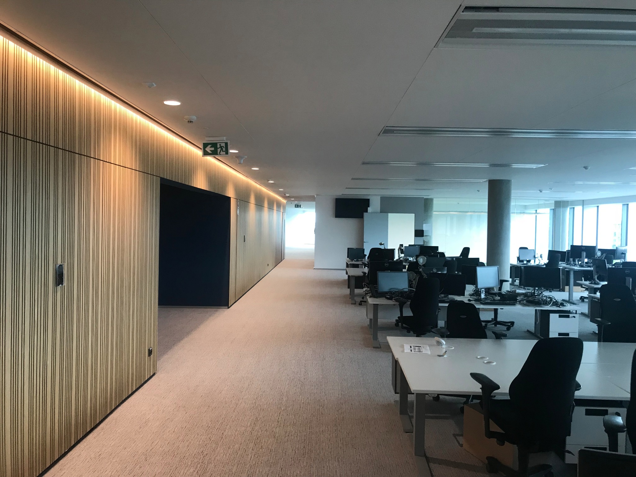 Wnętrze przestronnego biura z jasną wykładziną, drewnianą ścianą z oświetleniem LED i rzędem biurek z komputerami, widoczne znaki ewakuacyjne.