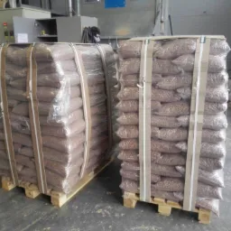 Dwie palety z workami brązowego pelletu owinięte folią i zabezpieczone taśmami transportowymi w magazynie.