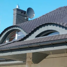 Szczegółowy widok dachu z gontu bitumicznego z dwoma półokrągłymi oknami dachowymi, ciemnymi rynnami i kominem z anteną satelitarną na tle błękitnego nieba.