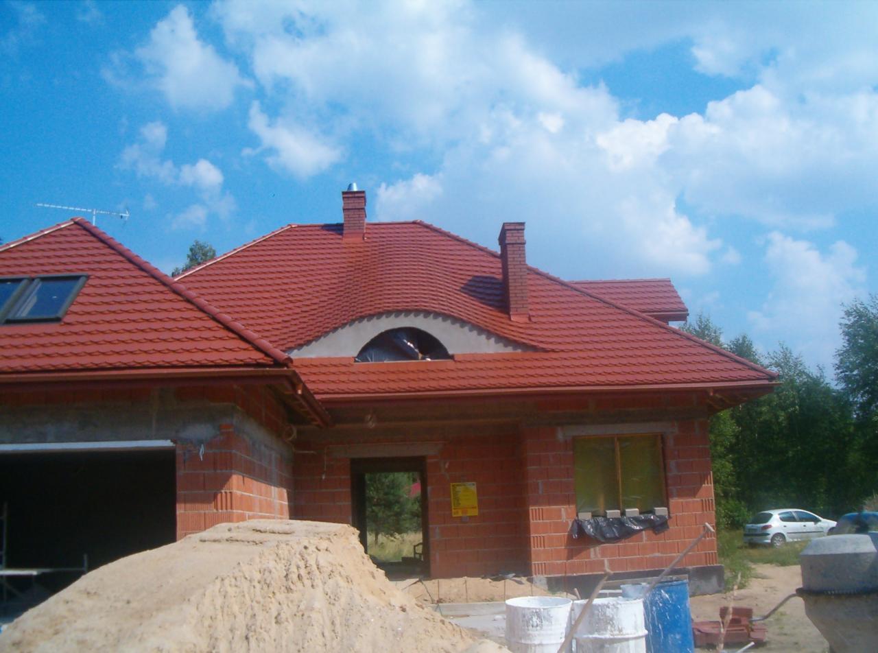 Nowo wybudowany dom z czerwoną dachówką, widoczne ceglane kominy i niezabudowane okno na poddaszu, na pierwszym planie sterta piasku i materiały budowlane.
