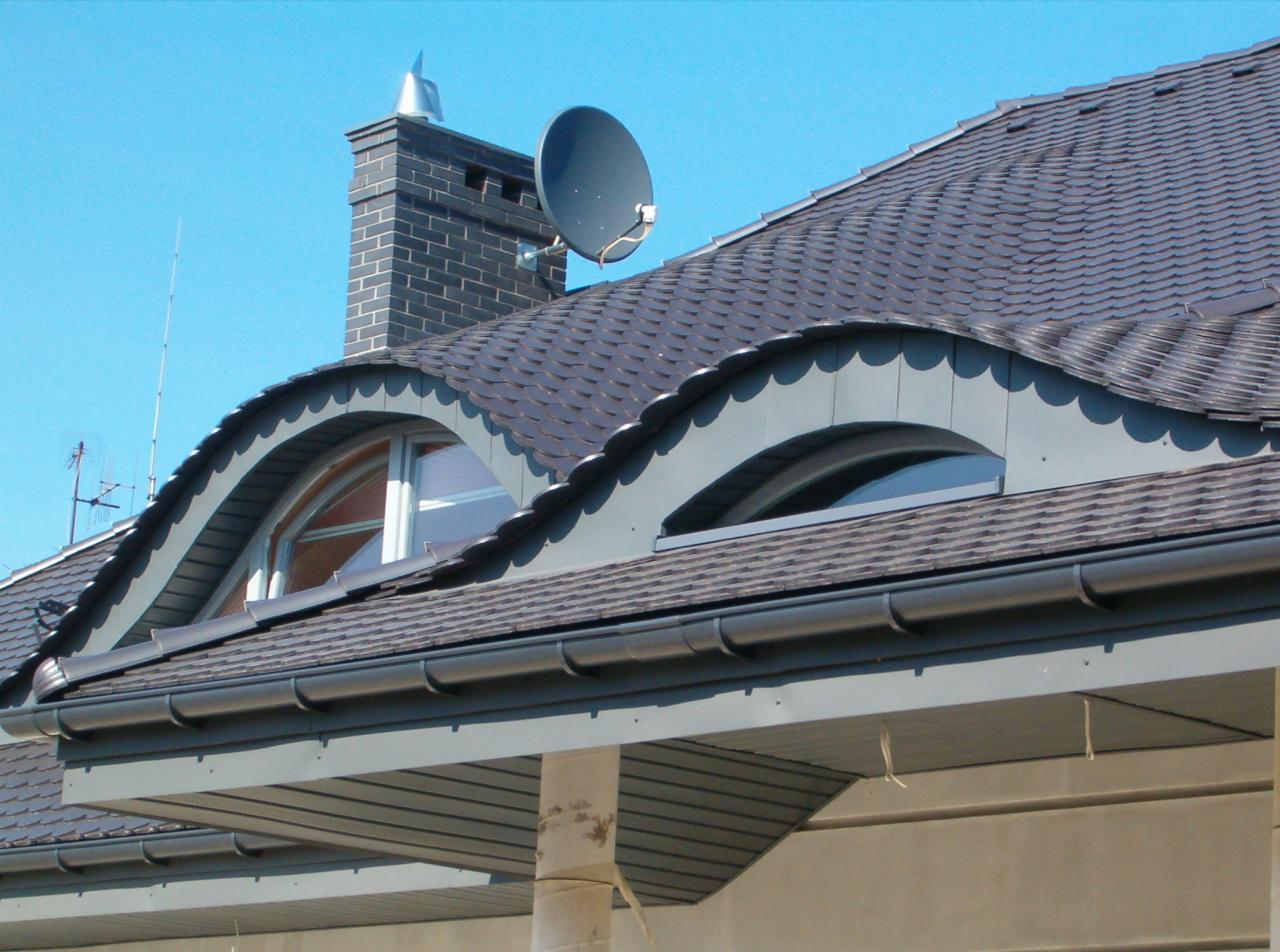 Szczegółowy widok dachu z gontu bitumicznego z dwoma półokrągłymi oknami dachowymi, ciemnymi rynnami i kominem z anteną satelitarną na tle błękitnego nieba.
