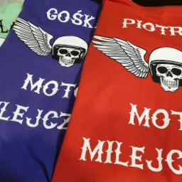 Kolorowe koszulki z nadrukiem czaszki w kasku z skrzydłami i napisem 'MOTO MILEJCZ' oraz imionami 'JUL', 'GOŚK' i 'PIOTREK'.