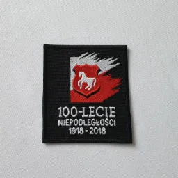 Czarna naszywka z wyhaftowanym motywem 100-lecia Niepodległości Polski (1918-2018), zawierająca stylizowany herb z białym koniem na czerwonym tle, ułożona na białym materiale.