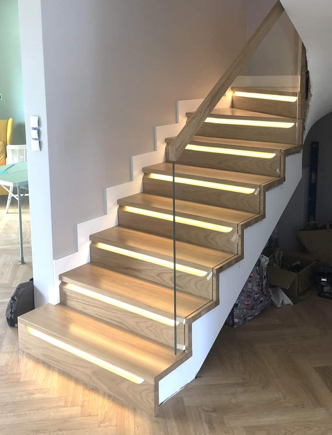 Nowoczesne schody z drewna z podświetleniem LED, balustrada ze szkła, widok z dołu na parkiet w jodełkę.