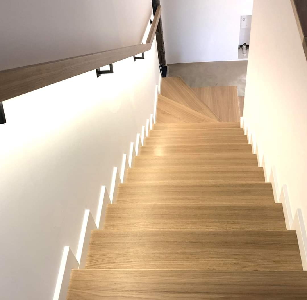 Jasne drewniane schody z podświetleniem LED wzdłuż ściany, z drewnianą poręczą zamocowaną na czarnych wspornikach, widok z góry.