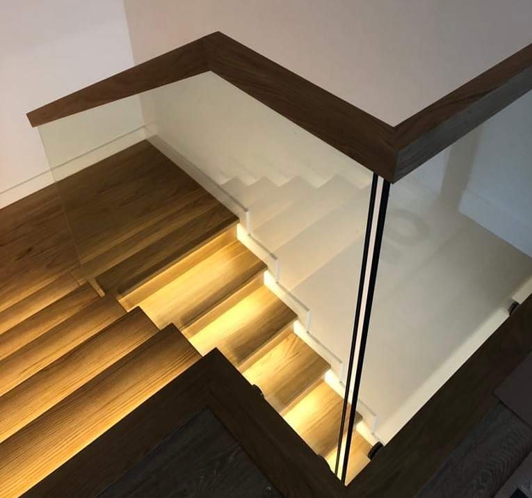 Nowoczesne schody z podświetlanymi stopniami, drewnianymi elementami wykończeniowymi i szklaną balustradą z czarnymi słupkami, widok z góry.