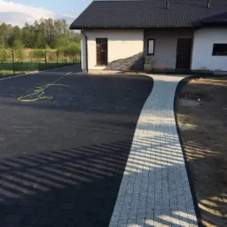 Nowoczesny dom z podjazdem wyłożonym ciemną kostką brukową i jasnym, granitowym chodnikiem prowadzącym do drzwi wejściowych, ogrodzenie panelowe w tle.