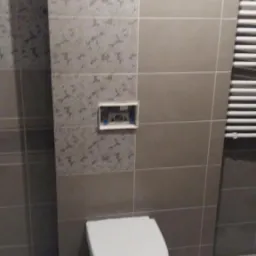 Ściana w łazience wyłożona szarymi i wzorzystymi płytkami ceramicznymi, z widocznym stelażem podtynkowym i podwieszaną toaletą.