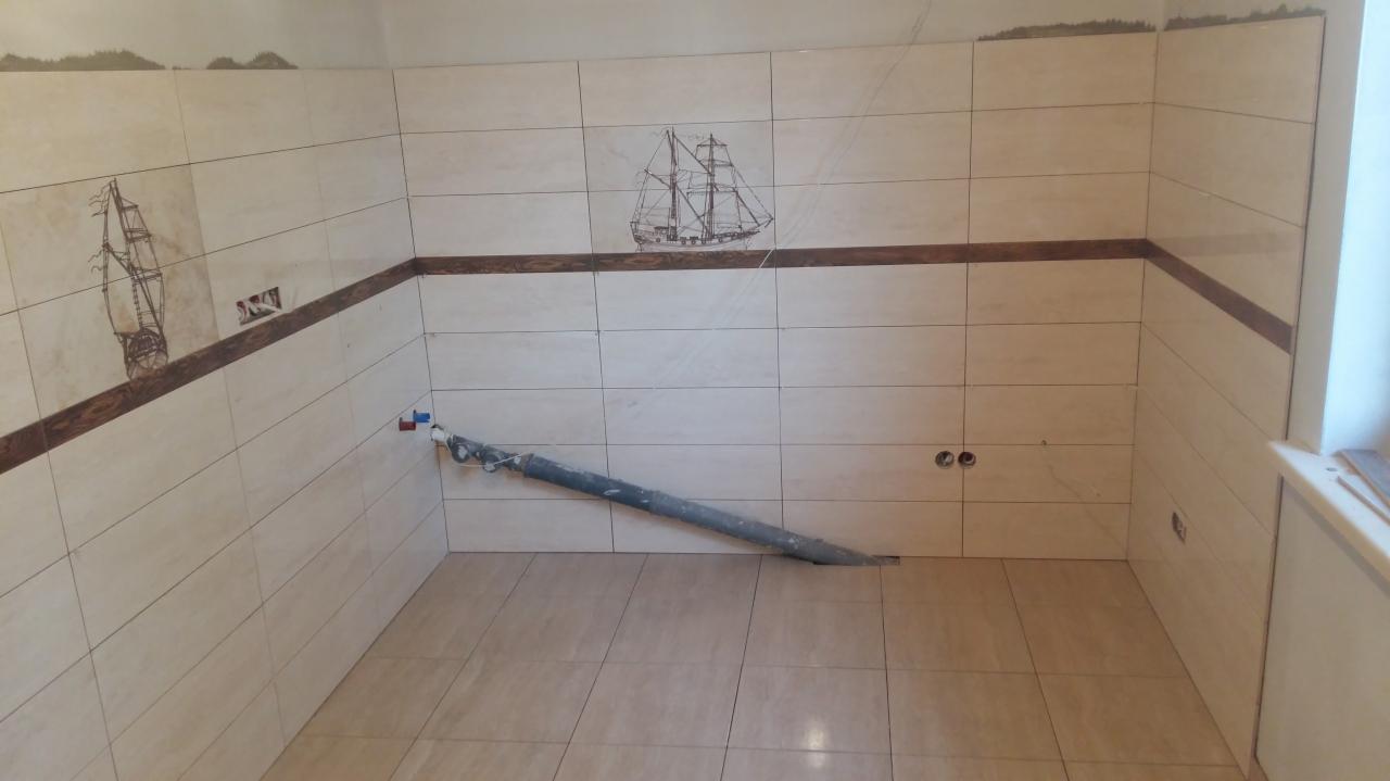 Ściany wyłożone jasnymi płytkami z dekoracyjnym motywem żaglowca i brązowym paskiem, widoczne rury instalacji wodnej i otwory na gniazdka elektryczne, pomieszczenie w trakcie remontu.