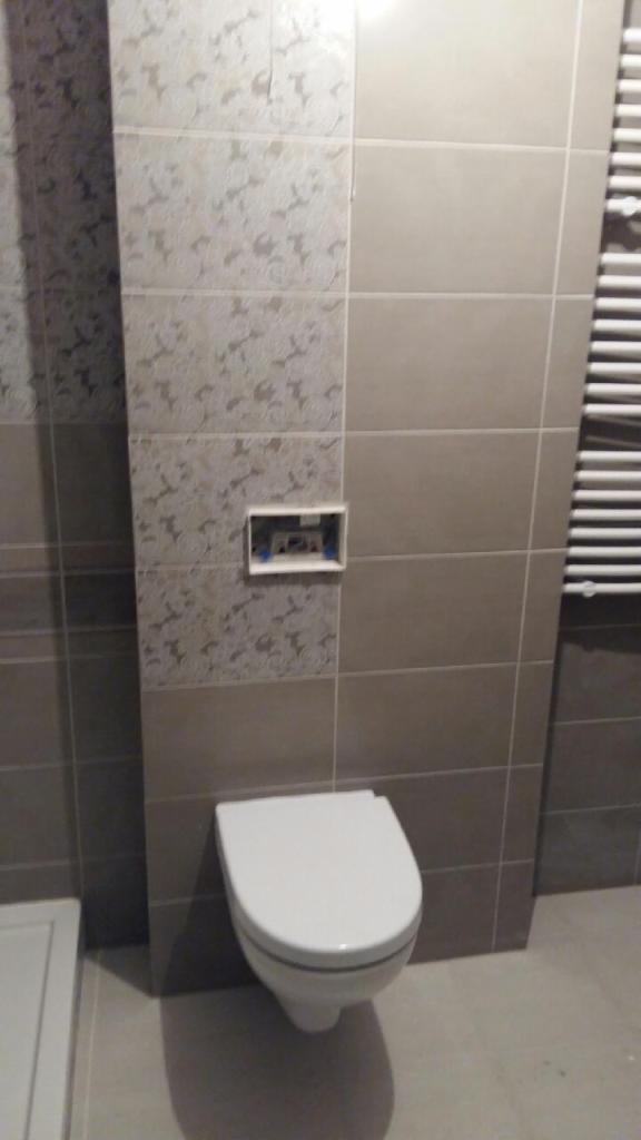 Ściana w łazience wyłożona szarymi i wzorzystymi płytkami ceramicznymi, z widocznym stelażem podtynkowym i podwieszaną toaletą.