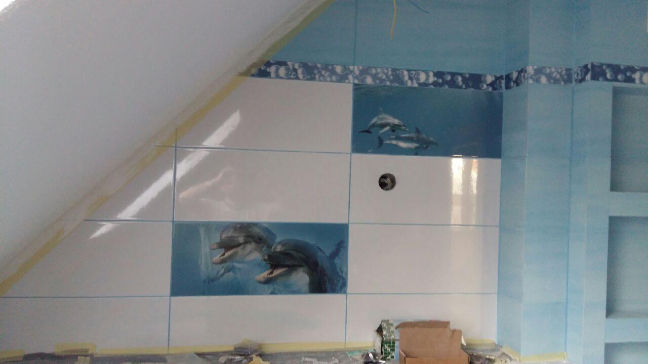 Ściana w łazience wyłożona białymi i błękitnymi płytkami z motywem delfinów, widoczny fragment skośnego sufitu z taśmą malarską, podłoga w trakcie prac wykończeniowych.