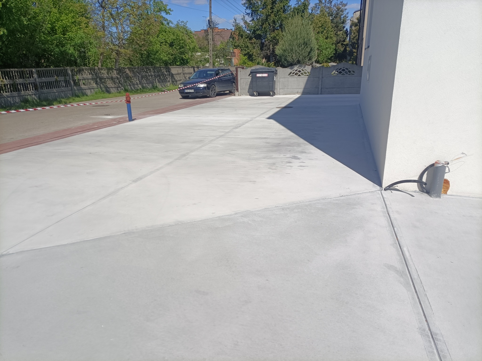 Świeżo wylana, gładka, szara betonowa posadzka na podjeździe przed domem jednorodzinnym, z widocznymi szczelinami dylatacyjnymi, zaparkowanym samochodem i pojemnikami na śmieci w tle.