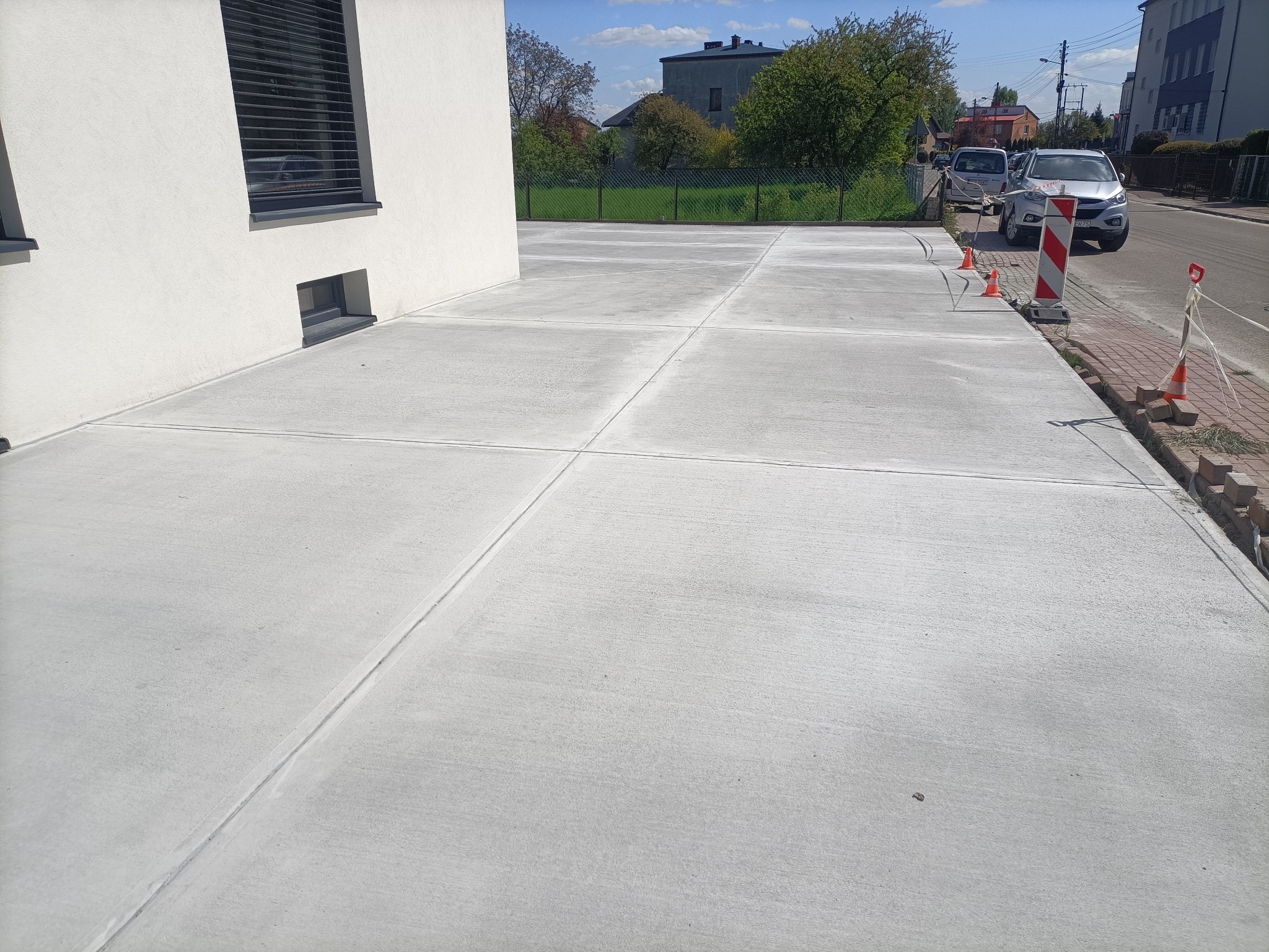 Świeżo wylana, podzielona na kwadraty, betonowa nawierzchnia przed budynkiem z widocznymi zabezpieczeniami w postaci słupków i pachołków drogowych.