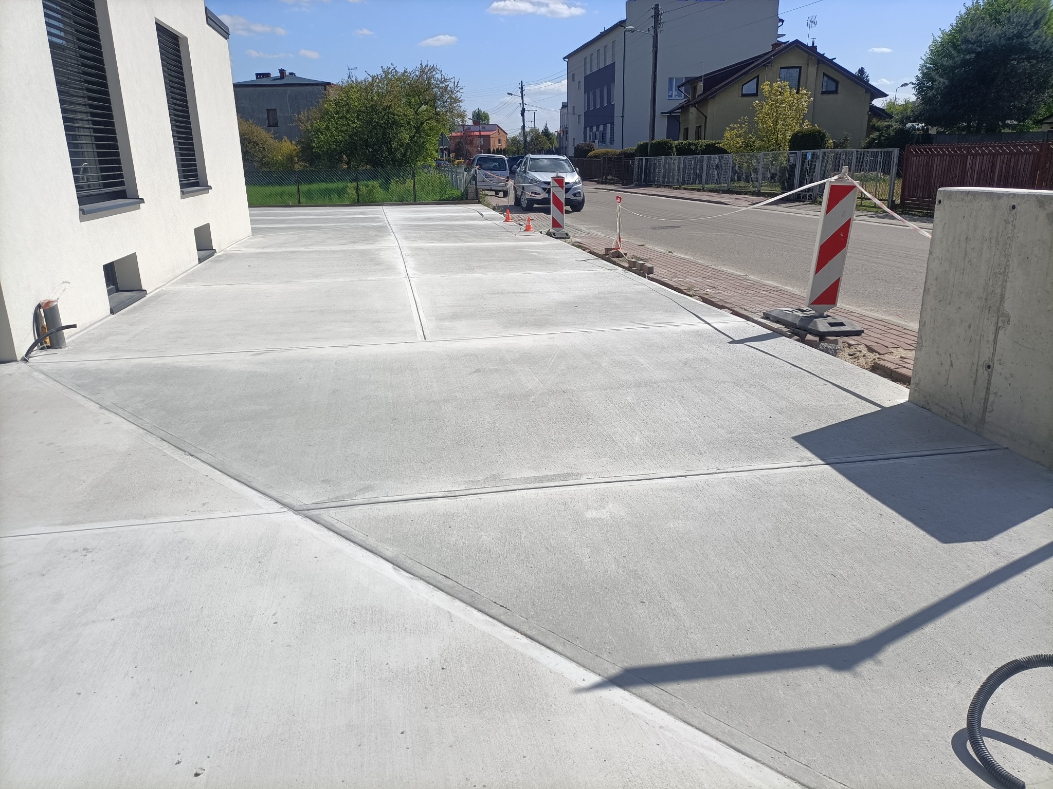 Nowa, jasnoszara powierzchnia betonowa podzielona na prostokątne segmenty, tworząca podjazd przed nowoczesnym budynkiem z ciemnymi oknami, zabezpieczona słupkami ostrzegawczymi i taśmą.