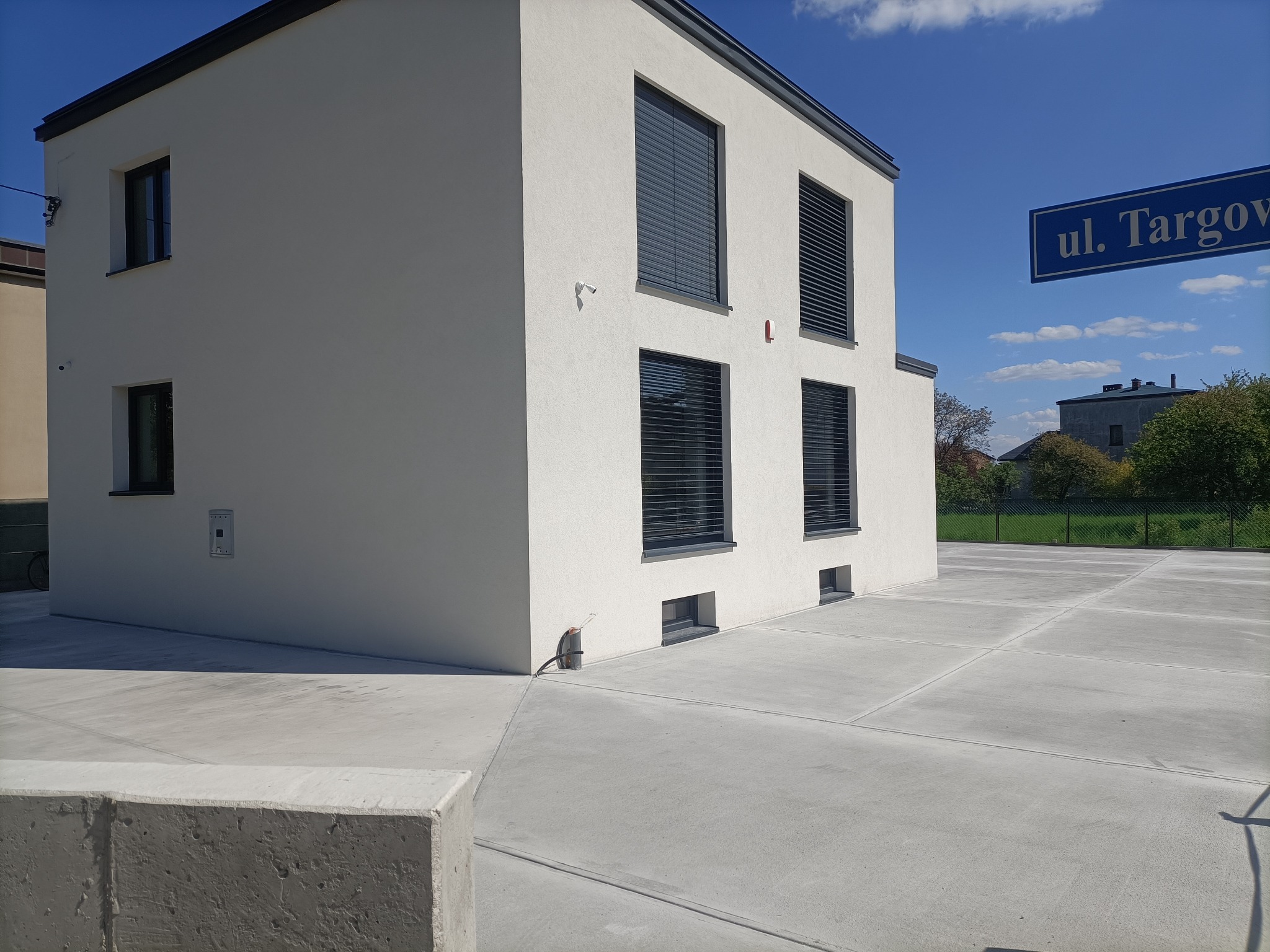 Nowoczesny budynek z jasną elewacją i szarymi ramami okiennymi, na zewnątrz betonowa posadzka podzielona na kwadraty, widoczny znak uliczny 'ul. Targowa'.