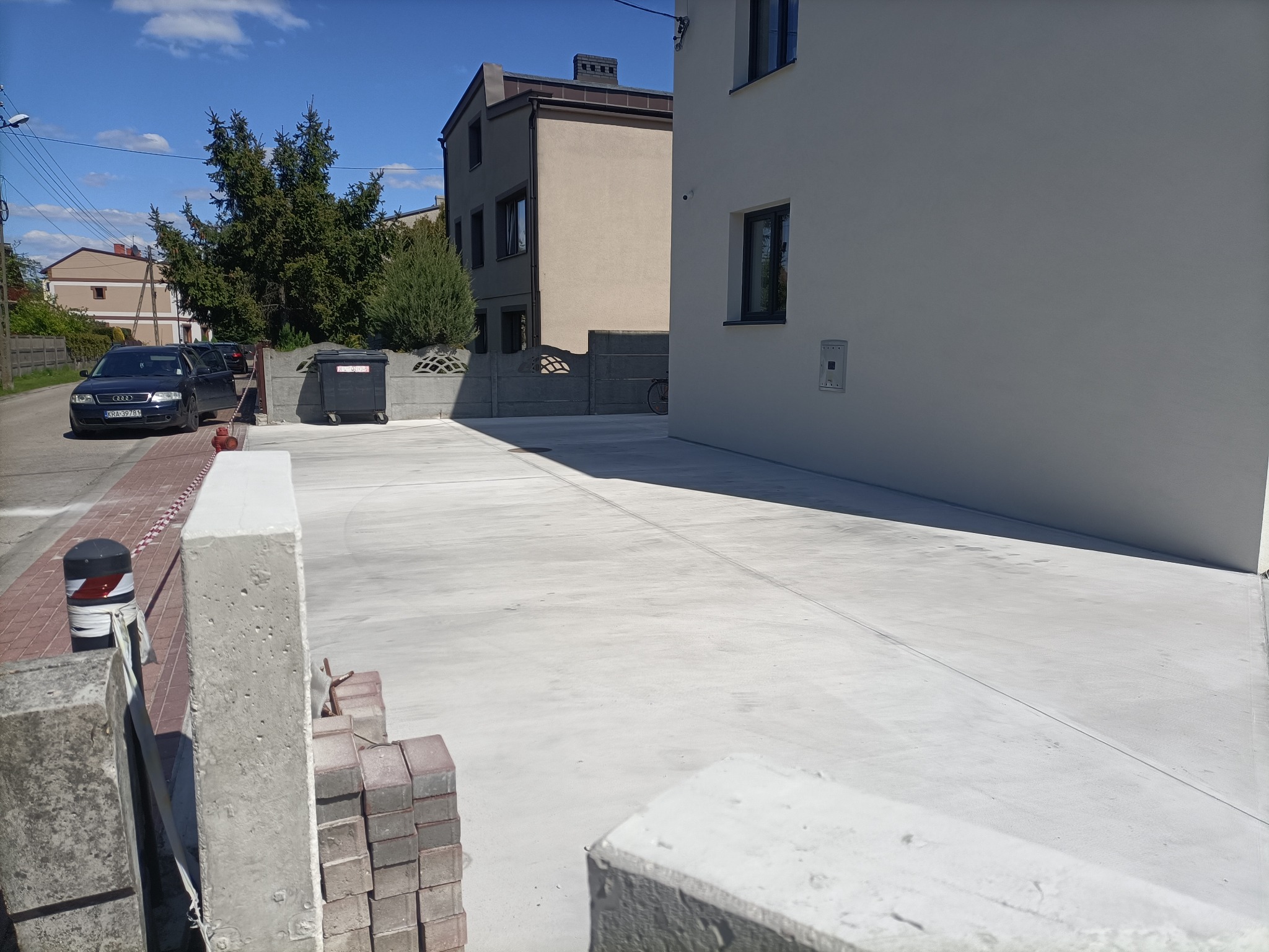 Świeżo wylana, jasnoszara posadzka na zewnątrz budynku, widoczne bloczki betonowe i kostka brukowa w pobliżu. W tle zaparkowany samochód osobowy.