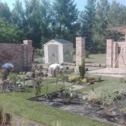 Układanie trawy z rolki i sadzenie roślin w ogrodzie z elementami architektury ogrodowej: ceglane słupki i drewniana konstrukcja altany w trakcie budowy.