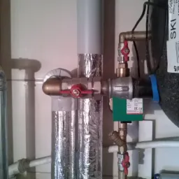 Instalacja centralnego ogrzewania z zaworami, rurami owiniętymi folią aluminiową i zieloną kostką sterującą, widoczne elementy podgrzewacza wody.