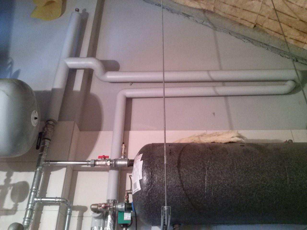 Szare rury instalacyjne, zbiorniki i zawory w piwnicy, widoczne elementy izolacji termicznej, oświetlenie fluorescencyjne.