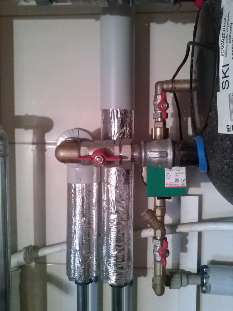 Instalacja centralnego ogrzewania z zaworami, rurami owiniętymi folią aluminiową i zieloną kostką sterującą, widoczne elementy podgrzewacza wody.