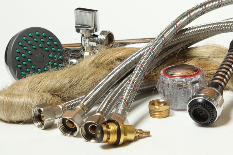 Zestaw hydrauliczny: główka prysznicowa z zielonymi dyszami, chromowany kran, węże w oplocie stalowym z czerwono-niebieskim wzorem, kurki, złączki mosiężne i pęk pakuł na białym tle.
