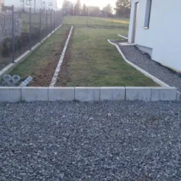 Ułożony obrzeże z betonowych elementów oddzielające trawnik od wysypanego kamieniami podwórka, widoczny fragment ogrodzenia i budynek w tle.