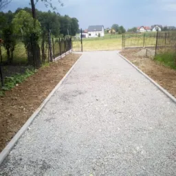 Utwardzona żwirowa ścieżka prowadząca do bramy z kutego żelaza, obrzeża z szarej kostki betonowej, w tle domy jednorodzinne i zieleń.