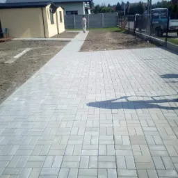 Ułożona szara kostka brukowa tworząca chodnik i podjazd, widoczny fragment ogrodu z domkiem narzędziowym w tle.