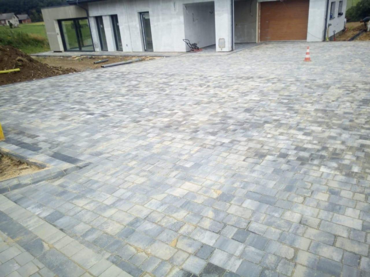 Ułożona szara kostka brukowa na podjeździe do nowoczesnego domu z bramą garażową, widoczny stożek ostrzegawczy i trawnik w tle.