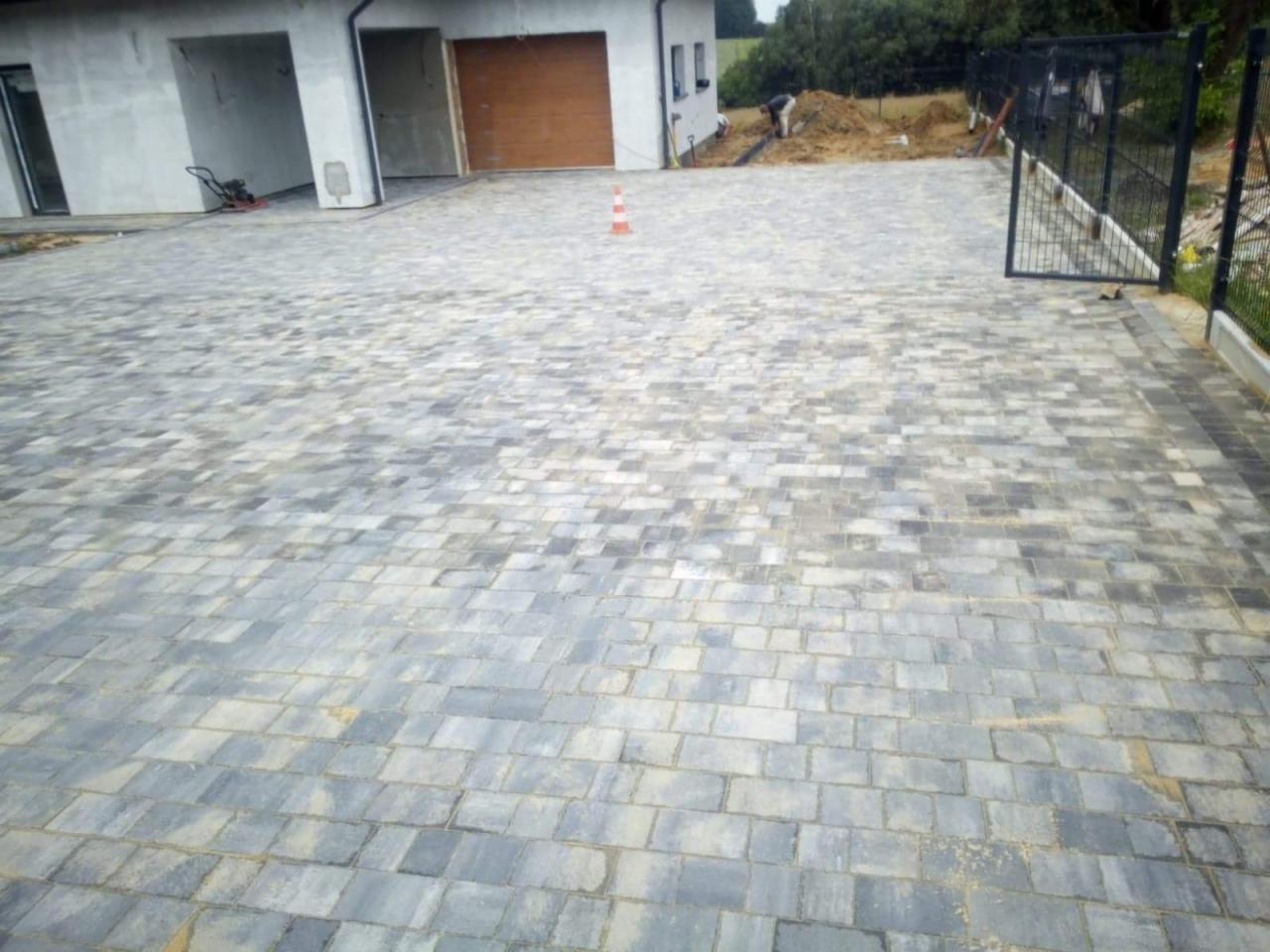 Ułożona szara kostka brukowa na podjeździe przed budynkiem z bramą garażową, widoczny fragment ogrodzenia i prace ziemne w tle.