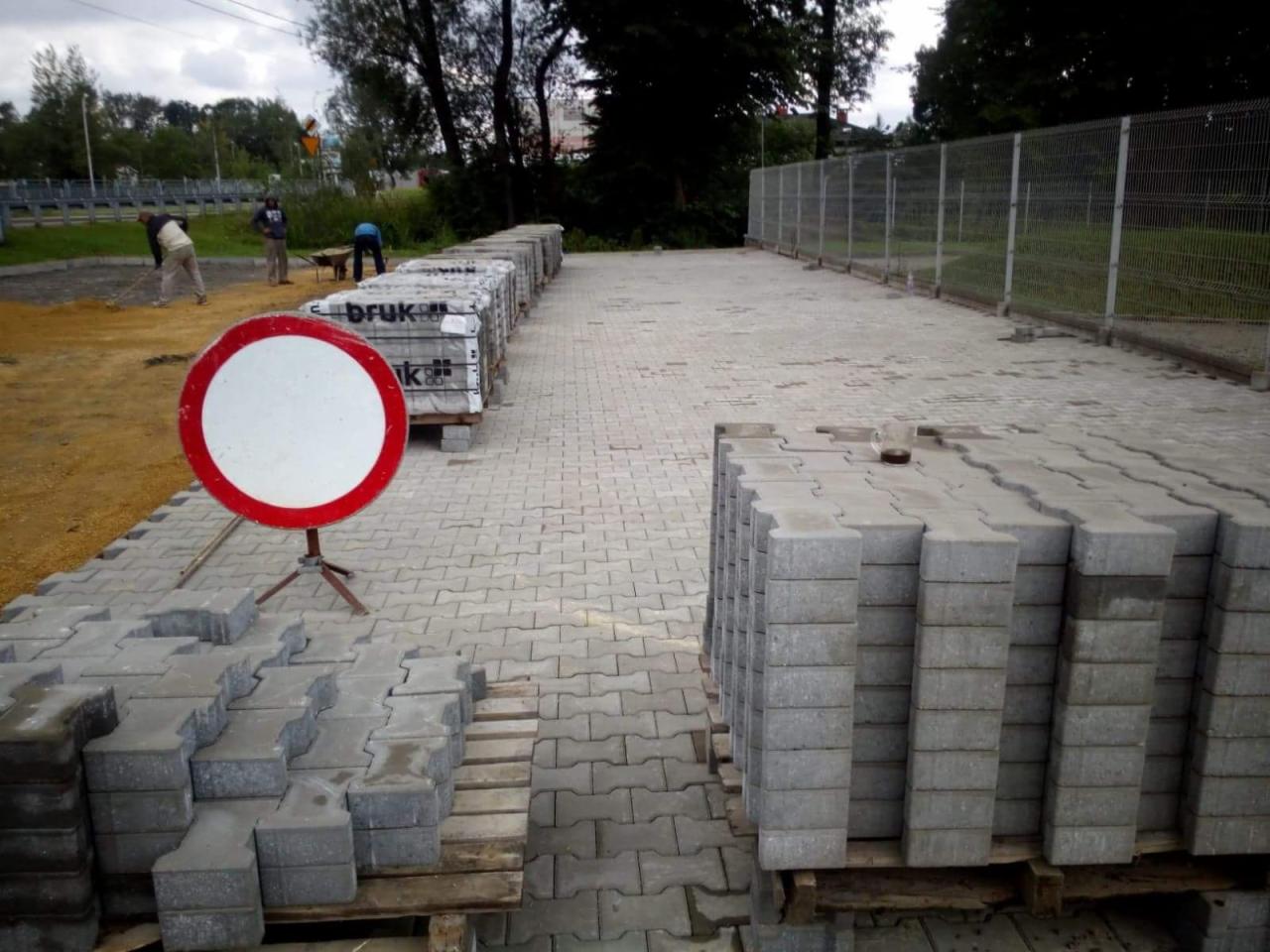 Plac budowy z ułożoną częścią szarej kostki brukowej, palety z kostką czekają na ułożenie, w tle robotnicy przygotowują podłoże, a obok stoi znak zakazu wjazdu.