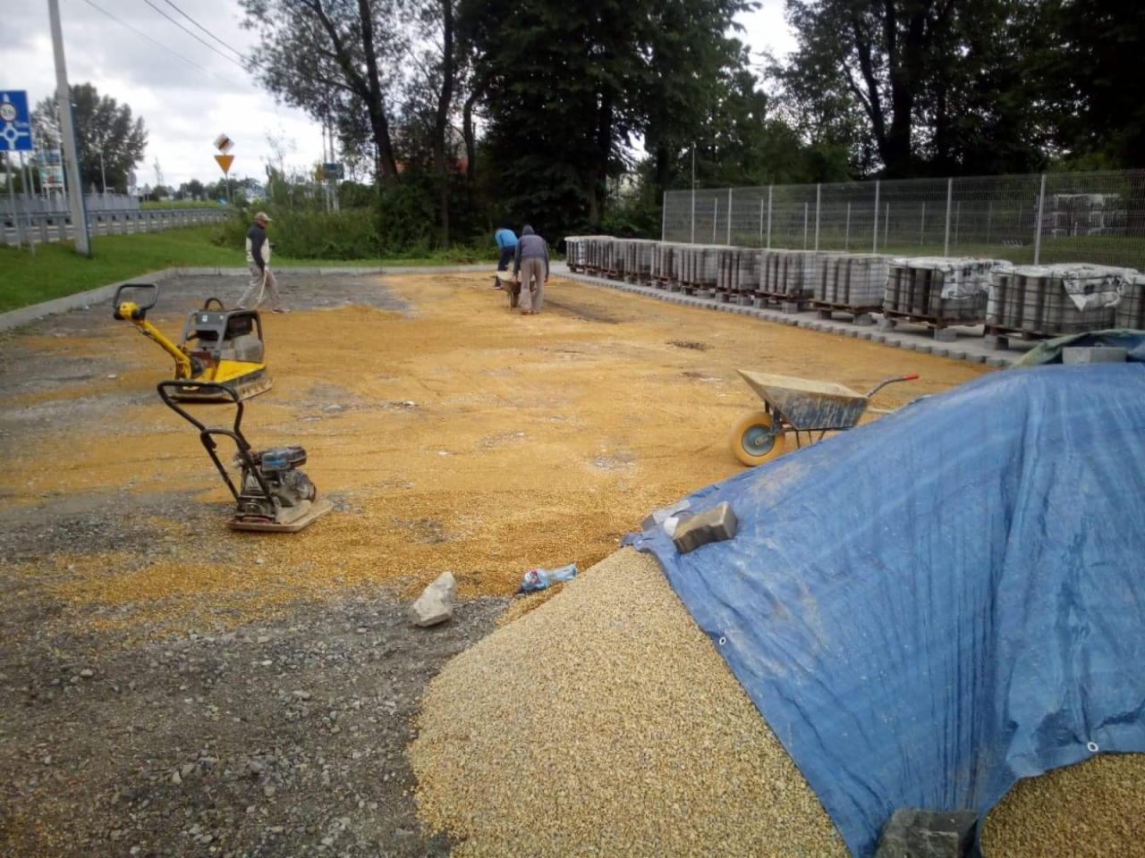 Prace brukarskie w trakcie: ubijarka wibrująca na podsypce, kostka ułożona w rzędach na paletach, taczka i brygada robotników w tle.