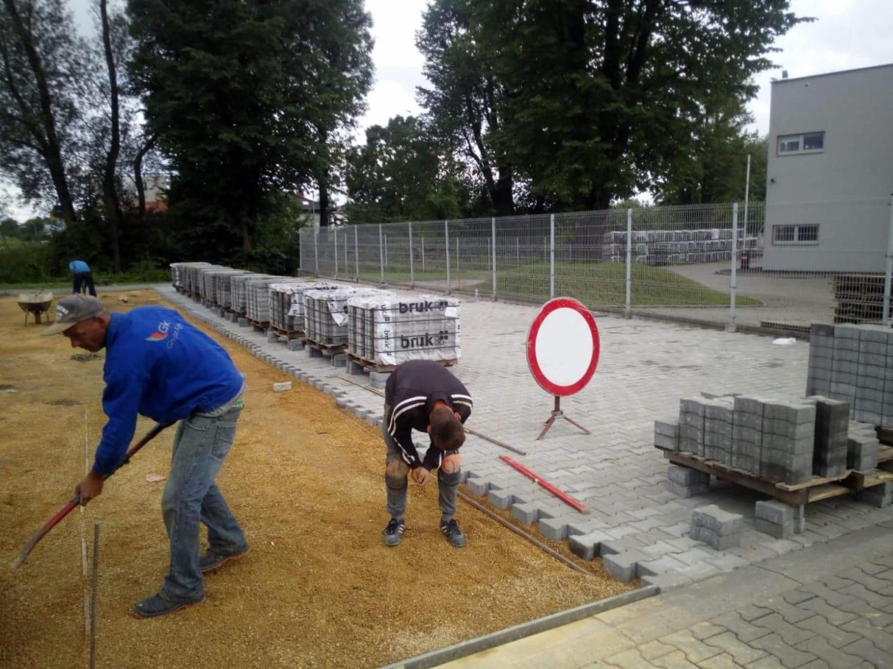 Dwóch pracowników układa szarą kostkę brukową na podsypce z piasku, palety z kostką w tle, znak zakazu wjazdu po prawej stronie.