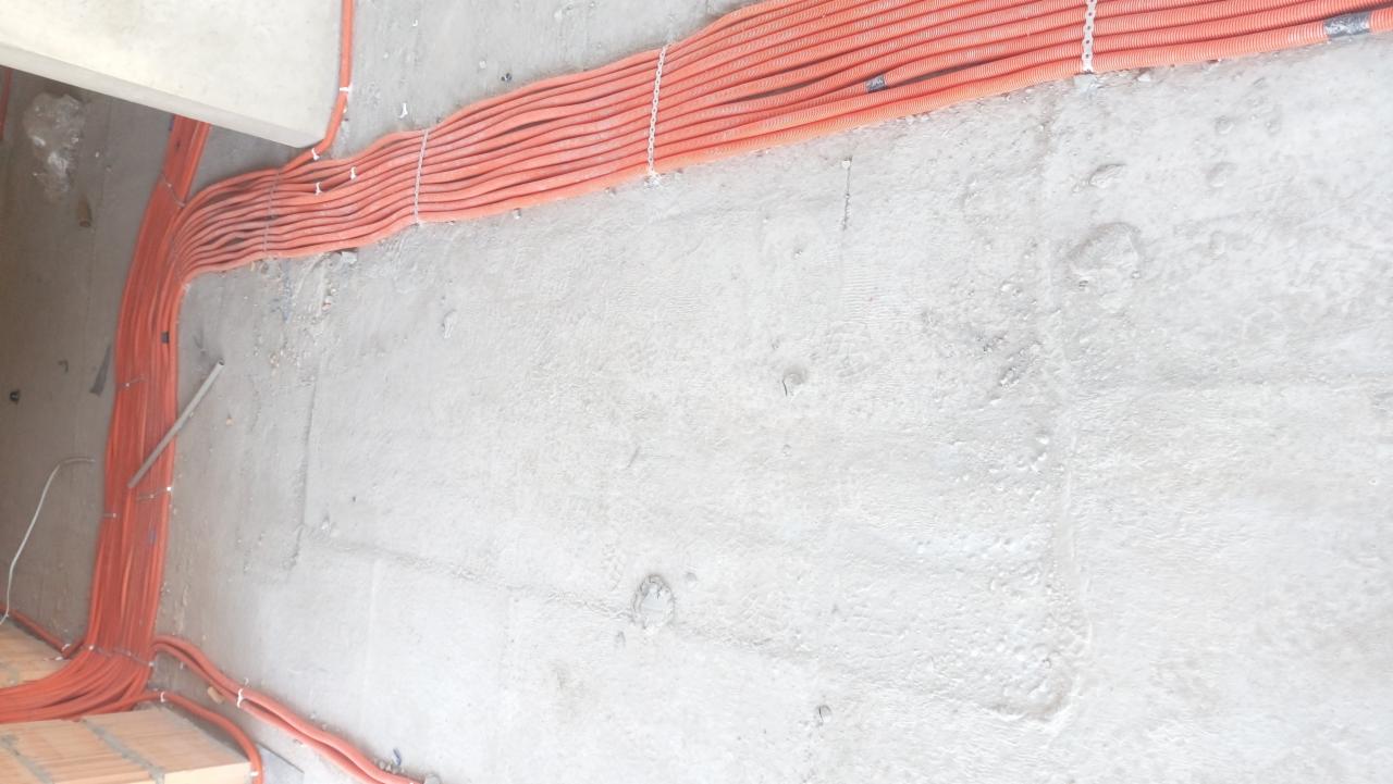 Wiązka pomarańczowych rur karbowanych zabezpieczonych białymi opaskami przymocowana do surowego, betonowego sufitu i ściany w nowym budynku.