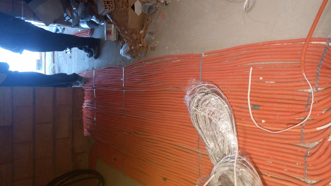 Układanie pomarańczowych rur instalacyjnych na ścianie z cegieł, widoczne kable w folii i fragment osoby w roboczych butach.