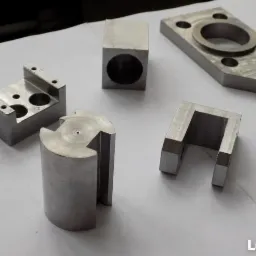 Zestaw precyzyjnie obrobionych, metalowych elementów o różnych kształtach, leżących na białym tle. Widoczne ślady obróbki skrawaniem.