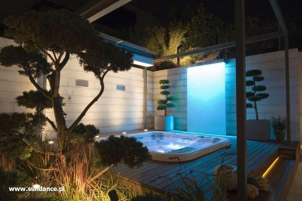 Nowoczesny ogród z podświetlanym jacuzzi, wodną ścianą i formowanymi drzewami w donicach, oświetlenie punktowe wkomponowane w roślinność.