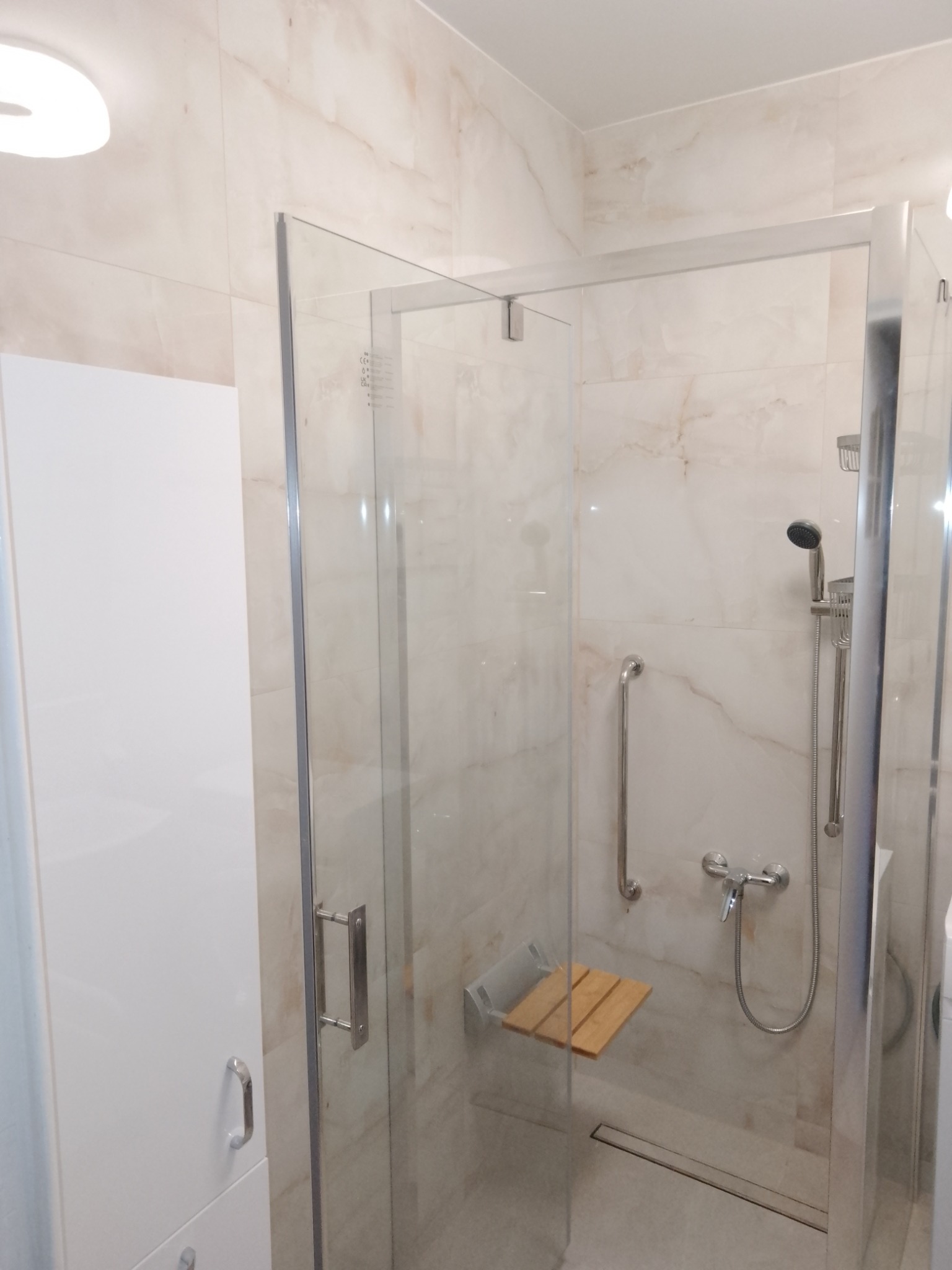 Nowoczesna kabina prysznicowa z siedziskiem z drewna, uchwytem dla niepełnosprawnych i armaturą w kolorze chromu, na tle beżowych płytek imitujących marmur.