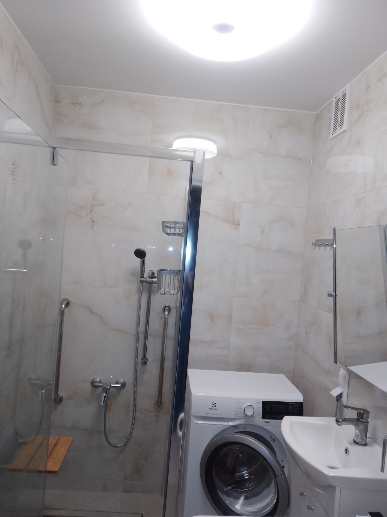 Wykończona łazienka z kabiną prysznicową ze szklanymi drzwiami, drewnianym siedziskiem wewnątrz, umywalką z szafką i pralką Electrolux pod jasnym oświetleniem sufitowym.