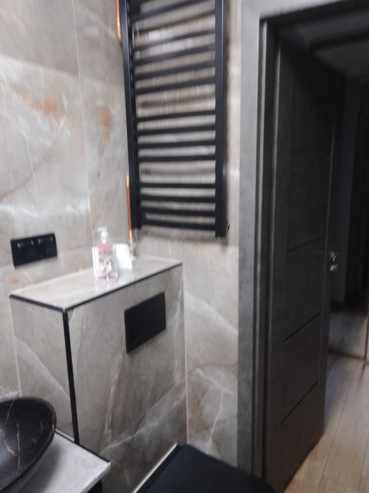 Nowoczesna łazienka z beżowymi płytkami imitującymi marmur, czarnym grzejnikiem drabinkowym, podwieszaną szafką z czarnymi elementami i widoczną umywalką w kolorze czarnego marmuru, oraz częściowo...