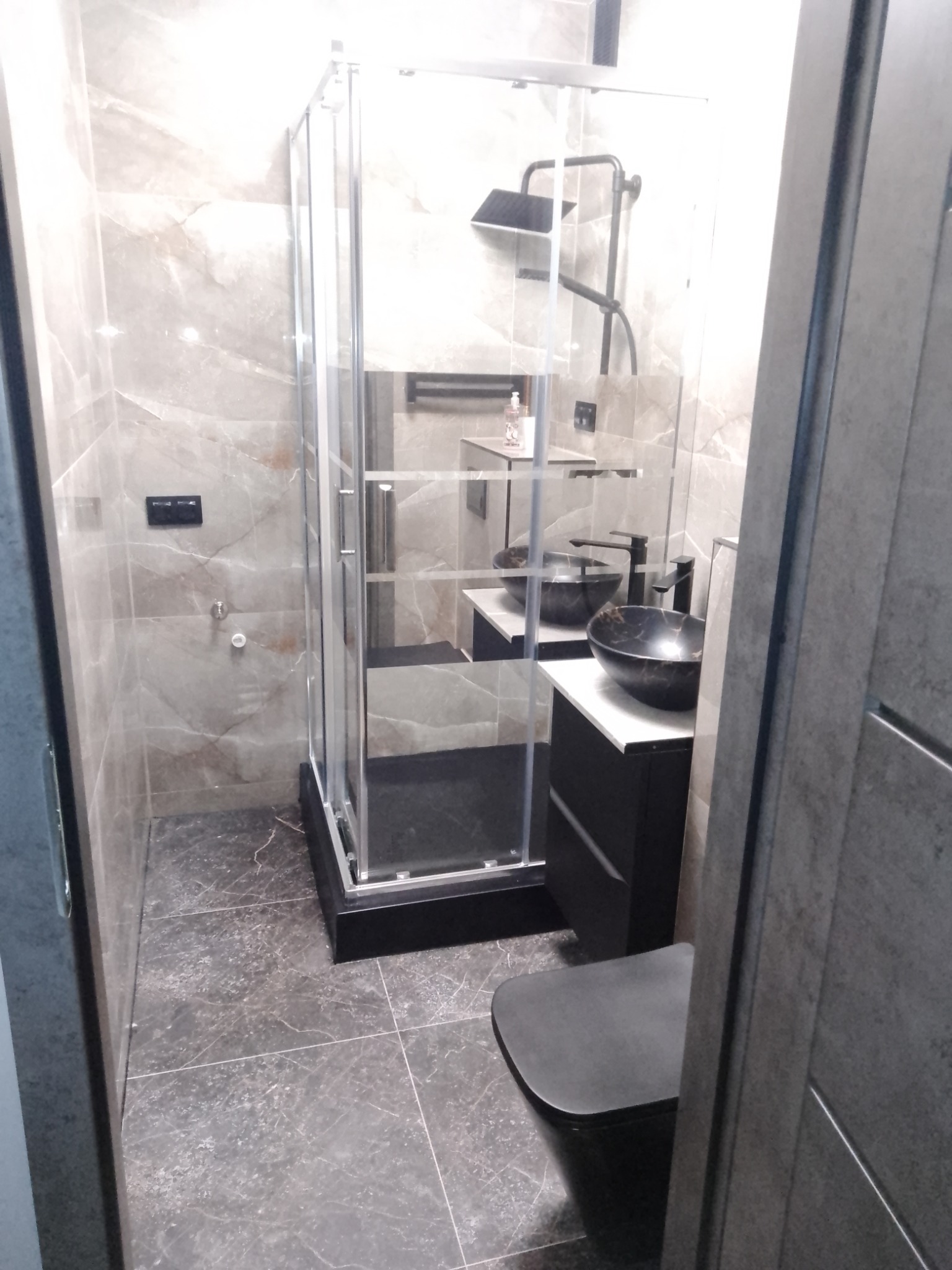 Nowoczesna łazienka z kabiną prysznicową, umywalką nablatową w odcieniach szarości i czerni, oraz toaletą w tym samym kolorze, wykończona marmurowymi płytkami.