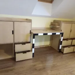 Pokój dziecięcy na poddaszu z biurkiem ze sklejki z motywem piłki nożnej i szafkami z szufladami, całość w jasnym drewnie.