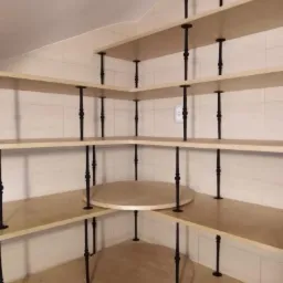 Zabudowa spiżarni z jasnej płyty stolarskiej z obrzeżem, mocowana na kutych, metalowych wspornikach, widok z wnętrza pomieszczenia na półki narożne i okrągłe.